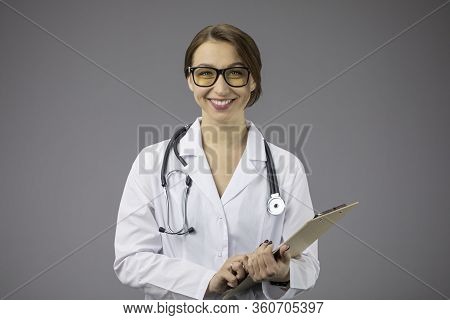 Smart Sexy Female Doctor In White Medical Coat And Stethoscope Holding Clipboard, Broadly Smiles, Fr