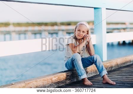 Portrait Of A Happy Little Girl Sitting On A Wooden Bridge With Sunlight