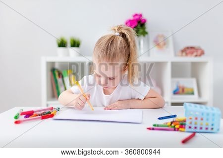 Blonde Happy Little Girl With Ponytale Drawing And Writing Lsitting By The White Table In Light Roon