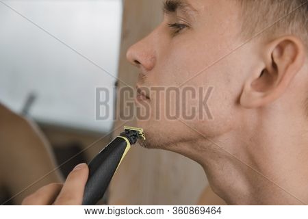 Close Up Of Young Man Looking To Mirror And Shaving Beard With Trimmer Or Electric Shaver