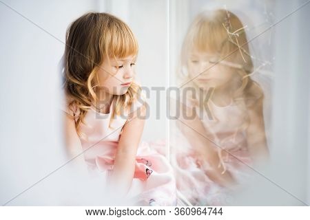 Sad Little Blonde Girl Looking Out Window. Child During Quarantine Because Of Pandemic Of Coronaviru