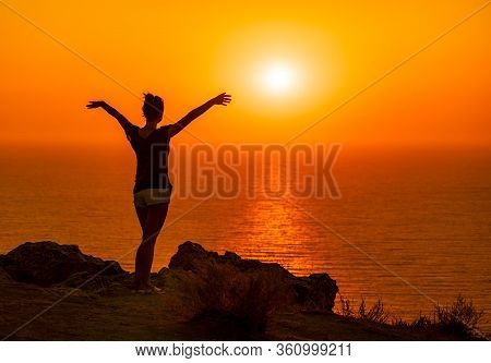 A Red Burning Sunset Over The Sea With The Silhouette Of A Young Woman Enjoying Evening