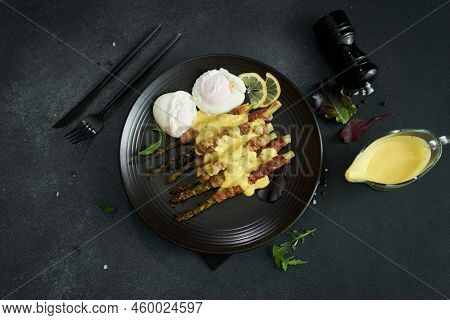 Baked Aspargus Covered With Bacon And Poached Eggs On A Black Ceramic Plate