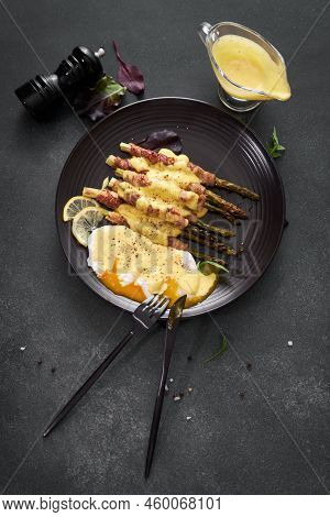 Baked Aspargus Covered With Bacon And Poached Eggs On A Black Ceramic Plate