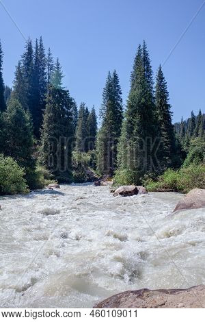 A Beautiful Stormy Mountain River Of Milky Hue Flows Rapidly Among Large Stones And Boulders Near Th