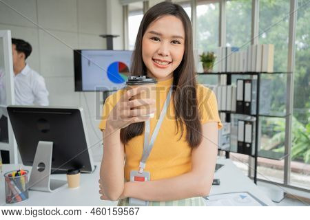 Young Asian Happy Businesswoman Or Intern Holding Coffice Cup In Modern Office Background Is Colleag