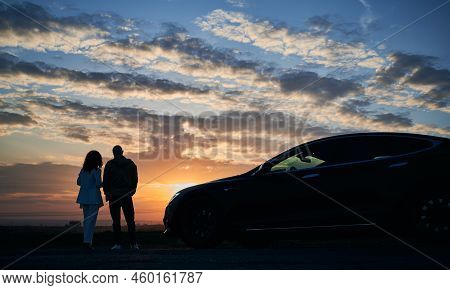 Silhouette Of Romantic Couple Who Arrived In Modern Car Out Of Town To Admiring Sunset Over Fields A