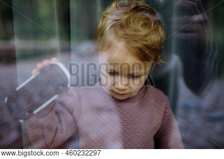 Little Child Standing Alone With Car Toy Behind The Window, Photo Trough Glass.