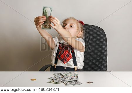 A Little Girl Sits At A Table In Front Of A Stack Of Hundred-dollar Bills And Examines One Of Them F