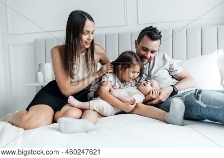 Young Parents And Their Children Play In Bed In A Bright Bedroom. Dad, Mom, Daughter And Newborn Bab
