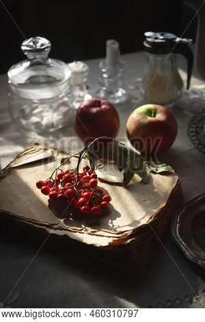 A Bunch Of Red Rowan Lying On The Old And Scrubby Book, Two Red Apples And The Other Tableware For T