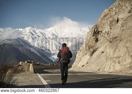 Rear View Of An Asian Hiker With Backpack Walking On Highway With Snowcapped Meili Mountain In Backg
