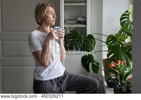 Tranquil Middle-aged Female Enjoys Hot Drink From Mug Standing At Windowsill. Mature Blonde Woman En