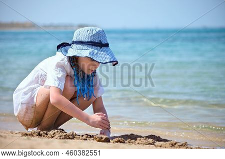 Happy Hild Girl In Big Hat And White Dress Playing Alone With Wet Sand On Sandy Beach Near Clear Sea