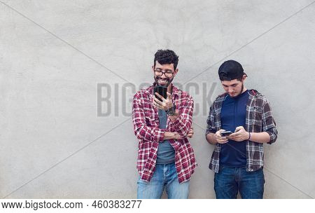 Two Smiling Friends Leaning On A Wall Checking Their Cell Phones, Friends Leaning On A Wall Texting