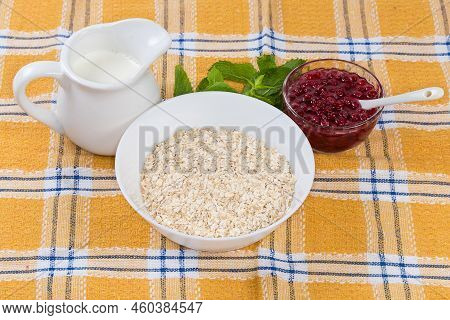 Raw Thin-rolled Oats In The White Bowl, Hot Milk In Milk Jug And Red Currants Jam On A Table With Ta