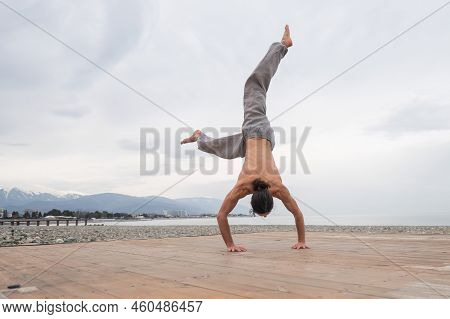 Shirtless Caucasian Man Doing Backflip On Pebble Beach.