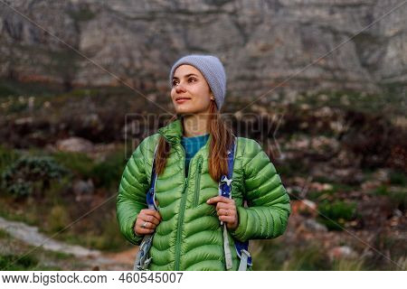 Thoughtful Young Female Hiker Wearing Down Jacket And Knit Hat Looking Away While Standing Against M