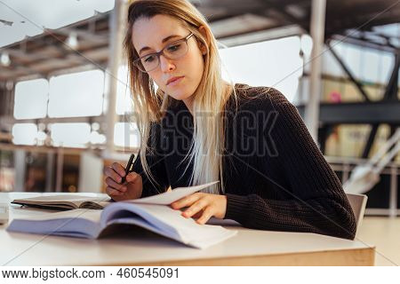 Focused Caucasian Young Woman With Blond Hair Wearing Eyeglasses And Studying On Desk In Library At 