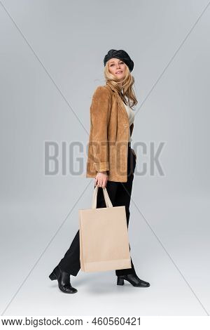 Full Length Of Blonde Model In Stylish Leather Beret And Beige Blazer Holding Shopping Bag On Grey.