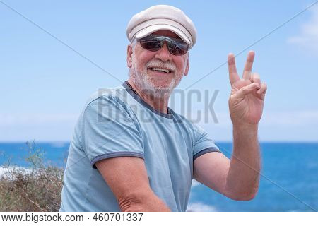 Smiling Caucasian Senior Man With Hat Sitting At The Beach Looking At Camera Gesturing Victory Sign 
