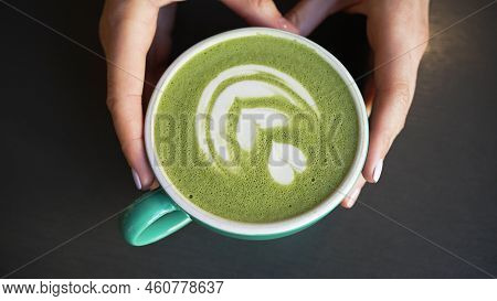 Female Hands Put Cup Of Green Matcha With Latte Art On Black Table. Barista Serves Matcha Latte In C