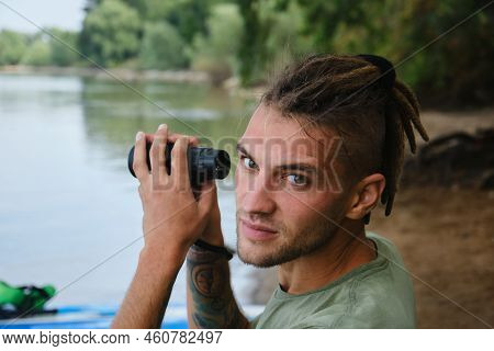 Caucasian Man With Dreadlocks Sits On Bank Of River In Summer And Holds Monocular In Hands. Device F