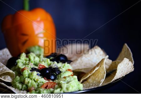 Black olive spiders crawling on guacamole with chips. Selective focus on bug with blurred foreground and background. Pepper as monster with cut out face carved into a Halloween pumpkin Jack O\\\'Lantern.