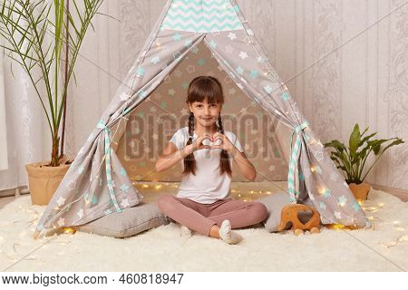 Horizontal Shot Of Lovely Kind Pretty Little Girl With Braids Wearing White T Shirt Posing In Wigwam