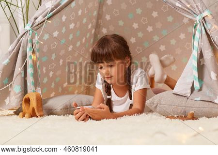 Dark Haired Little Girl With Braids Wearing White T Shirt Posing In Peetee Tent, Lying With Cell Pho