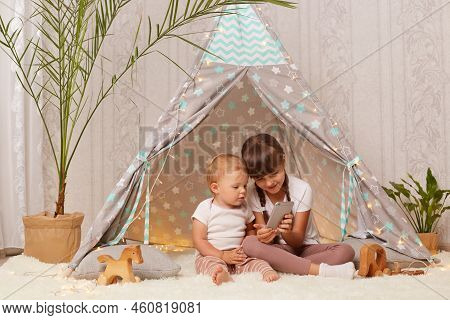 Indoor Shot Of Cute Positive Happy Sibling Girls Wearing White T Shirts Sitting In Peetee Tent And U
