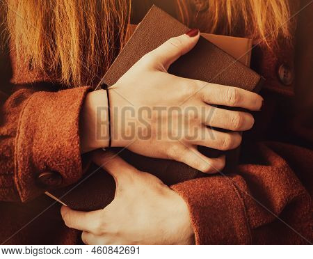 A Woman With Ginger Hair, Dressed In A Brown Woolen Jacket, Holds A Stack Of Old Books In A Hard Bro