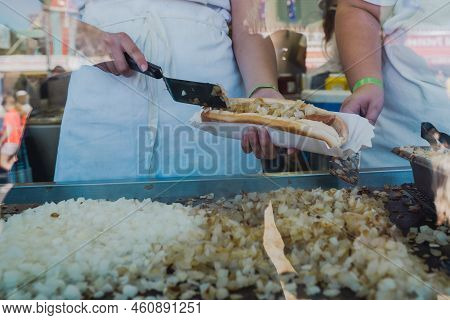 Worker Scoops Grilled Onions Onto A Footlong Hotdog While Working In A Booth At A Fair