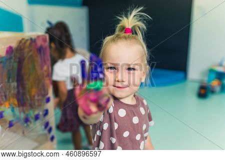 Cling Film Painting At The Nursery School. Happy Smiling Blonde Toddler Girl Showing Her Painted Col