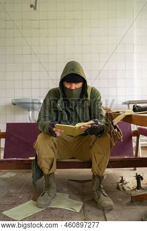 Man In Bandana Mask, Hood And Camo Pants Sits Inside Of Abandoned Building And Reading Documents. Po