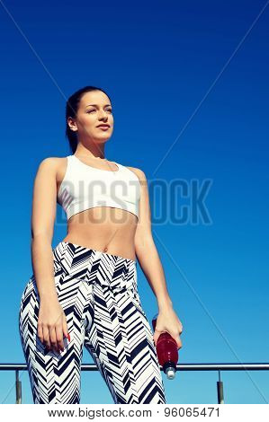 Young athletic female standing on blue sky background while holding bottle of energy drink