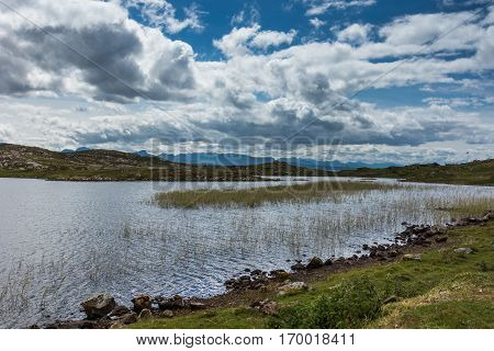 Assynt Peninsula Scotland - June 7 2012: Loch Sgeireach is a lake along B869. Here under blue sky with large white clouds. Reed in water some grass upfront. Horizon of mountains.