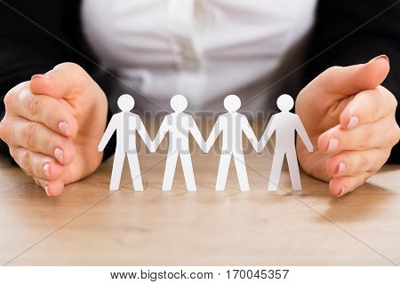 Close-up Of A Businesswoman Protecting Cutout Of Human Figure On Wooden Desk