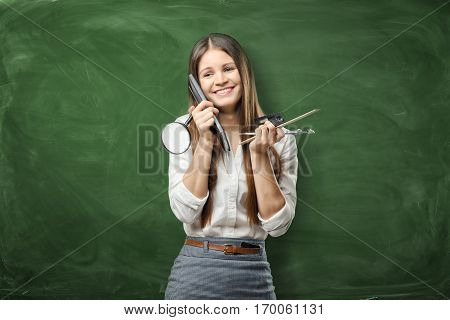 A smiley businesswoman standing and holding different office supplies in her hands, on the green background. Daily planning. Time-management. Secretary work.