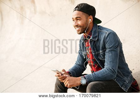 Cheerful african young man wearing cap walking on the beach and chatting by his phone while listening music with earphones.