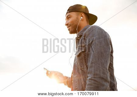 Photo of cheerful african young man wearing cap walking on the beach and chatting by his phone while listening music with earphones.