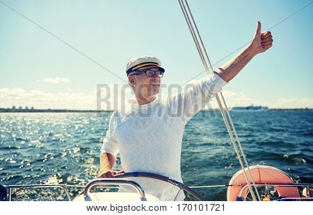 sailing, age, tourism, travel and people concept - happy senior man in captain hat on steering wheel and showing thumbs up sail boat or yacht floating in sea