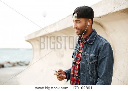 Cheerful african man wearing cap walking on the beach and chatting by his phone while listening music with earphones. Look aside.