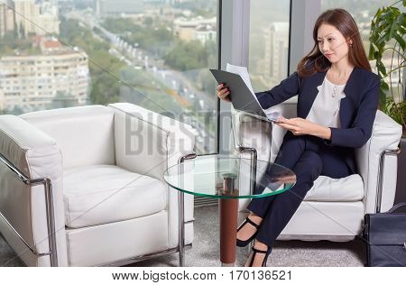 Young asian business woman in suit looking at the chart on the sheet sitting in an easy chair in the office
