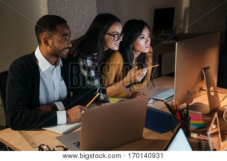 Photo of happy business people working late at night in their office with laptops and computers. Woman pointing with pencil to computer.