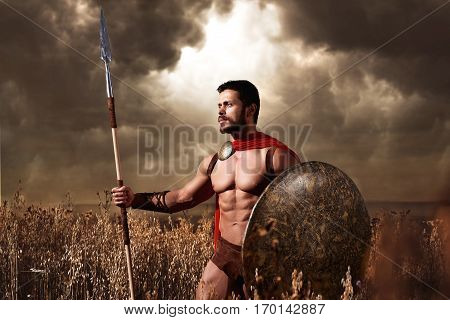 Athletic warrior with beard wearing like spartan looking away and holding iron weapon in hand. Brunet soldier with bare torso in red cloack going in attack with rounded shield and weapon. War in field.