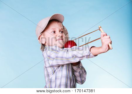 Nature and free time prankster little boy. Child have fun with wooden sling shot and fruit aim apple.