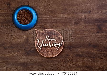 Bowl of dry kibble dog food. Mmm yummy speech bubble. Healthy pets feed. Blue plate on wooden rustic background.