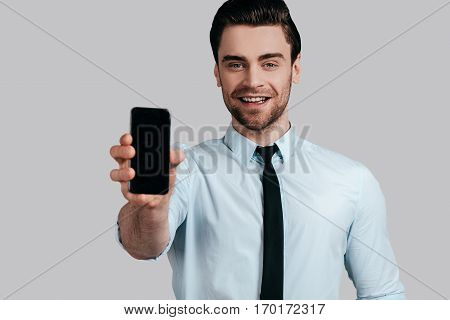 Copy space on his mobile phone. Good looking young man in white shirt and tie showing a smart phone and looking at camera while standing against grey background