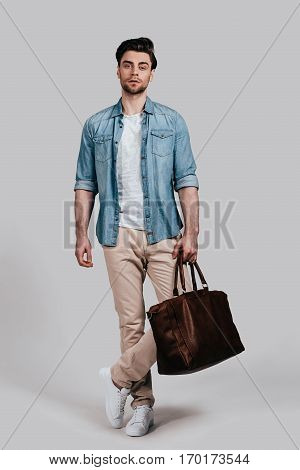 Stylish handsome man. Full length of confident young man in casual wear carrying brown leather bag and looking at camera while standing against grey background
